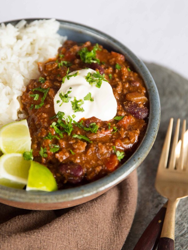 chilli served in small bowl with rice, sour cream and lime wedges next to golden spoon and fork