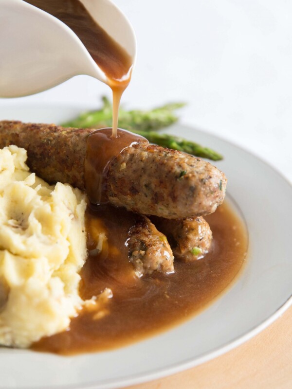 pouring gravy over sausages on white plate with asparagus and mash