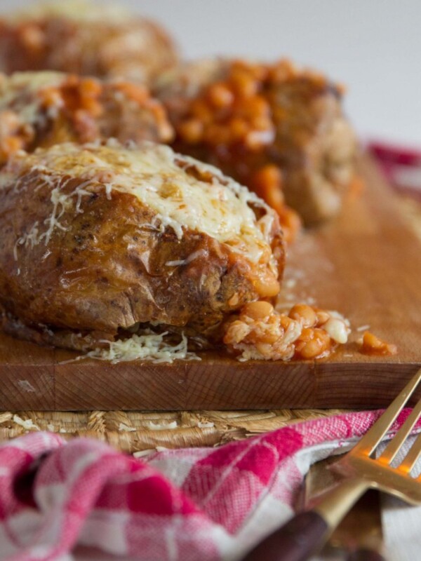 baked potato with beans pouring out on chopping board with 3 more blurred in the background