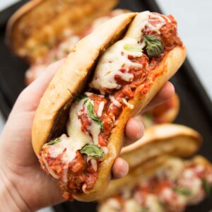 closeup shot of a hand holding meatball sub with melted cheese and 3 subs in the background