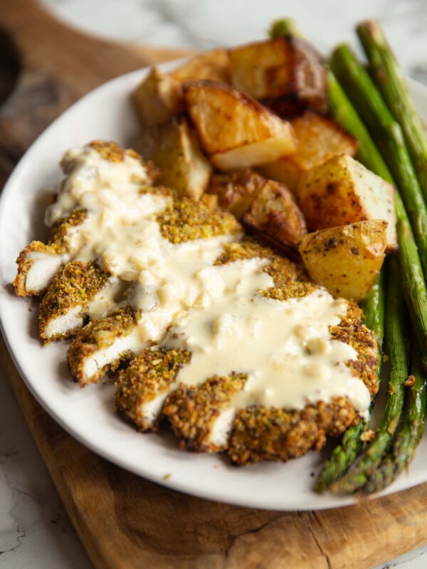 pistachio crusted chicken served on small white plate with potatoes and asparagus with creamy sauce on top