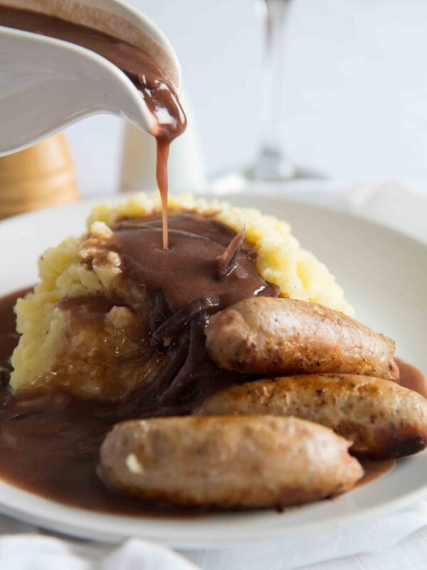 gravy being poured over mashed potato with sausages on small white plate