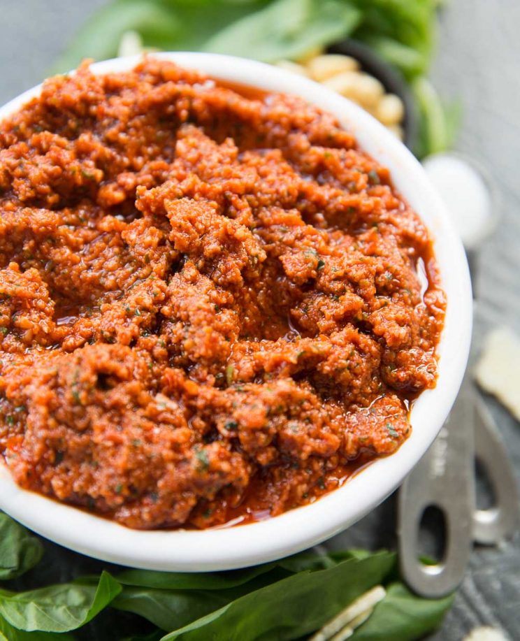 Sun Dried Tomato Pesto in a white bowl surrounded by fresh basil and pine nuts