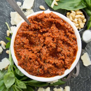 Bowl of Sun Dried Tomato Pesto with spoon poking out surrounded by basil, pinenuts and parmesan