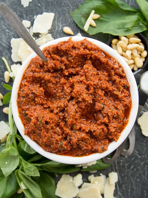 Bowl of Sun Dried Tomato Pesto with spoon poking out surrounded by basil, pinenuts and parmesan