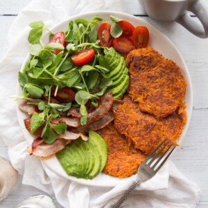 Sweet Potato Hash Browns on a white place with a side salad, bacon and avocados. Coffee and salt and pepper in the background.