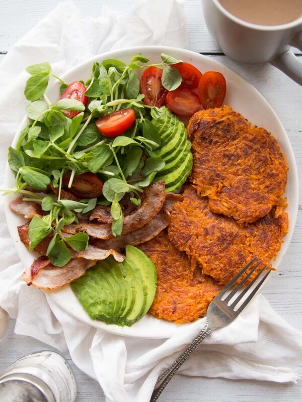 Sweet Potato Hash Browns on a white place with a side salad, bacon and avocados. Coffee and salt and pepper in the background.