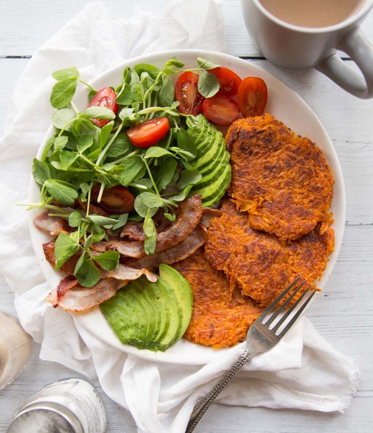 Hash Browns on a white place with a side salad, bacon and avocados. Coffee and salt and pepper in the background.