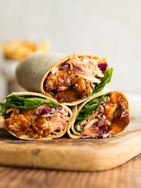 3 bbq chicken wraps stacked on a wooden chopping board with french fries blurred in the background