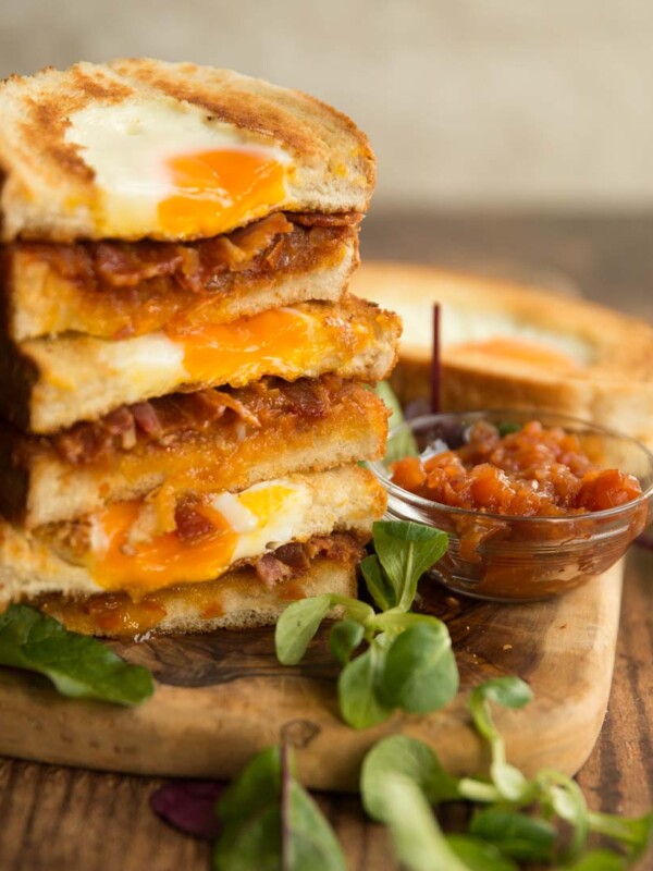 egg and bacon sandwiches stacked on top of each other on wooden board with salad to garnish