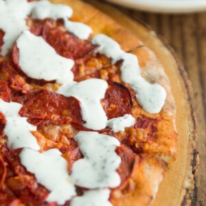 garlic and herb dip drizzled over pepperoni pizza on wooden board with potato wedges blurred in background