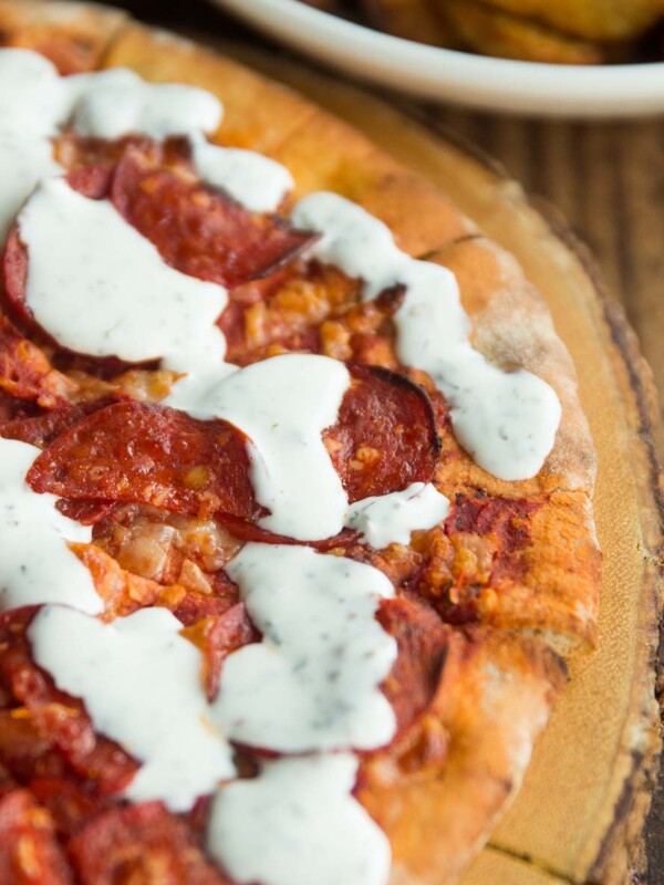 garlic and herb dip drizzled over pepperoni pizza on wooden board with potato wedges blurred in background