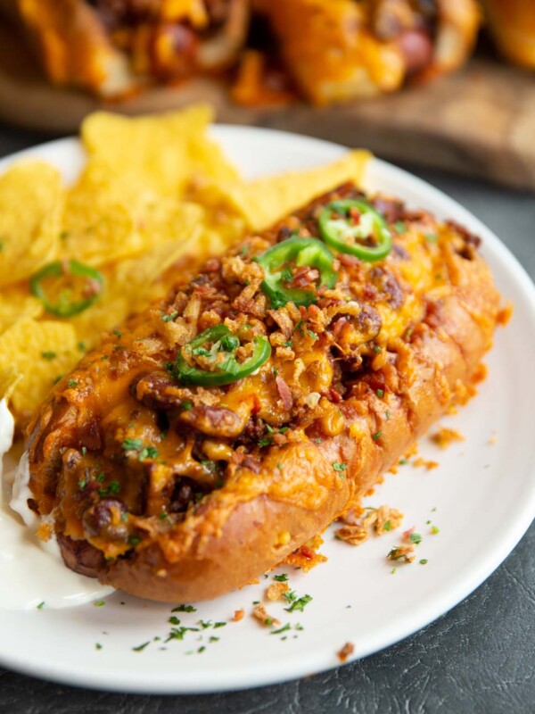 chili dog on white plate served with tortilla chips and more chili dogs blurred in background