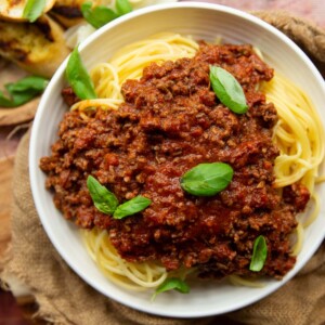 overhead shot of bolognese on spaghetti in white bowl with garlic bread blurred in corner