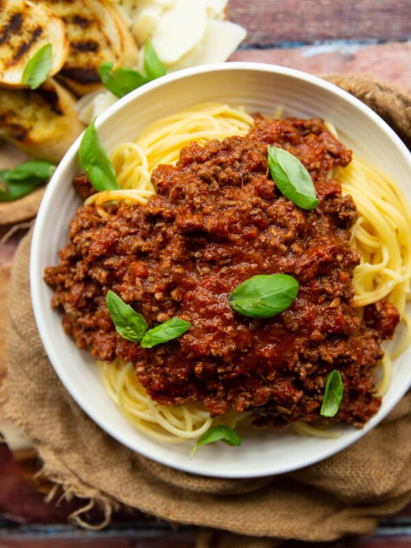 overhead shot of bolognese on spaghetti in white bowl with garlic bread blurred in corner