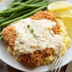 sauce drizzled over schnitzel on mash potato with green beans and lemon wedges blurred in background