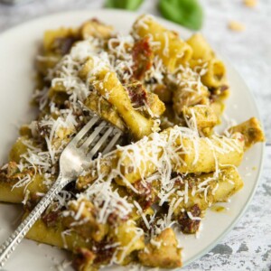 silver fork digging into pasta on white plate with basil and pine nuts blurred in background