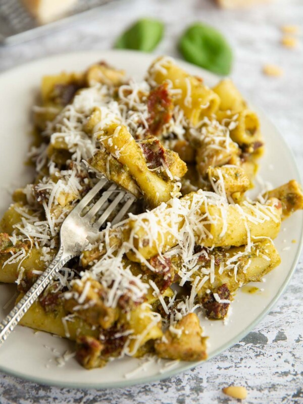 silver fork digging into pasta on white plate with basil and pine nuts blurred in background