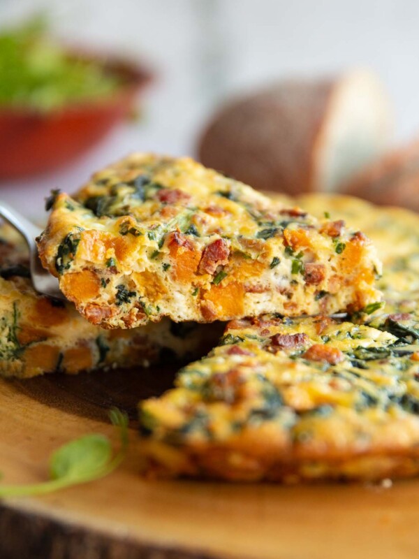 lifting slice of frittata above wooden board with salad and bread blurred in background