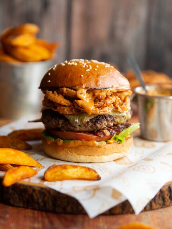 cowboy burger on shees of paper on wooden board surrounded by potato wedges