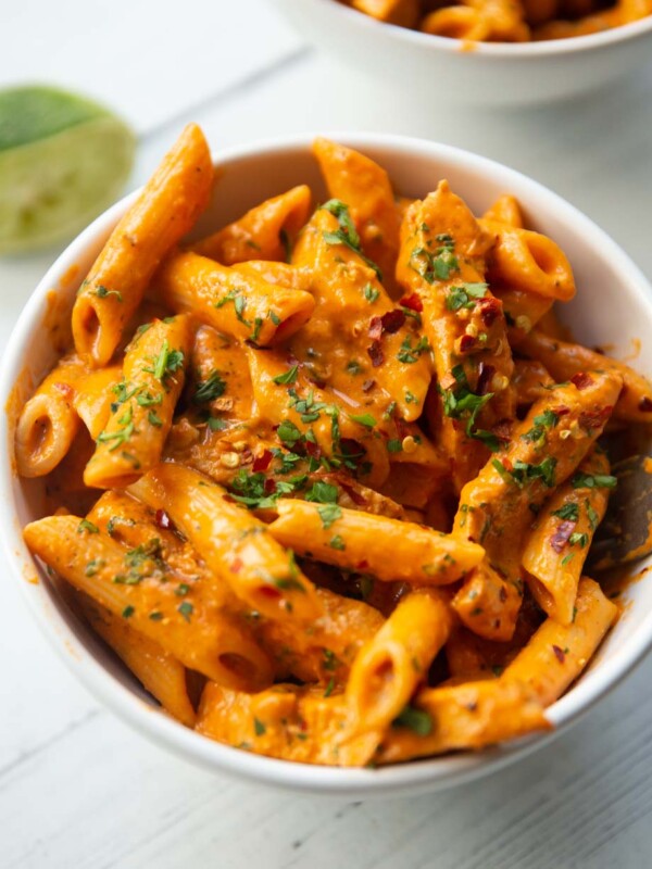 close up shot of pasta in small white bowl garnished with chilli flakes and parsley with lime blurred in background