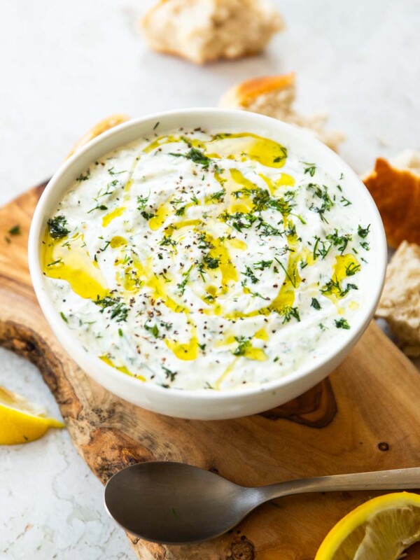 Tzatziki served in small white bowl on wooden chopping board surrounded by bread and lemon