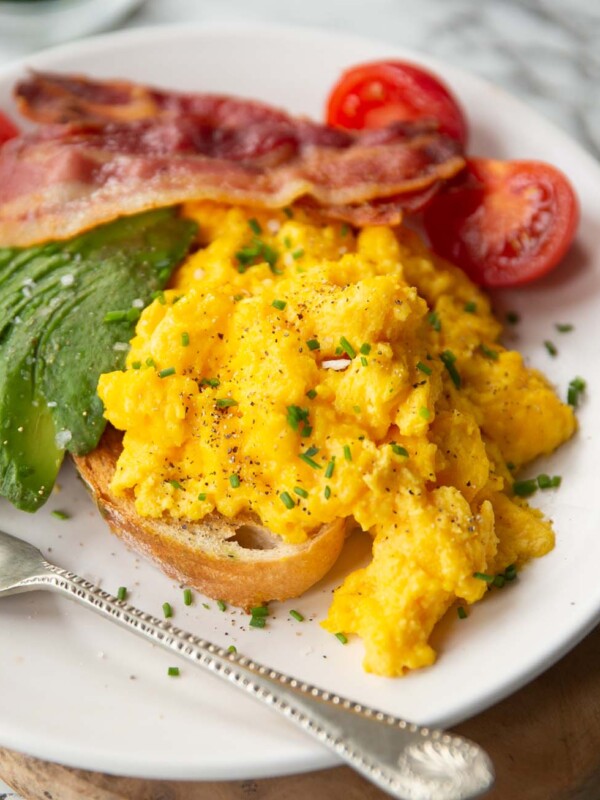 cheesy scrambled eggs served on toast on white plate with bacon, tomatoes and avocado