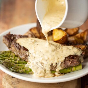 close up shot of small white pot pouring sauce over steak with potatoes and asparagus