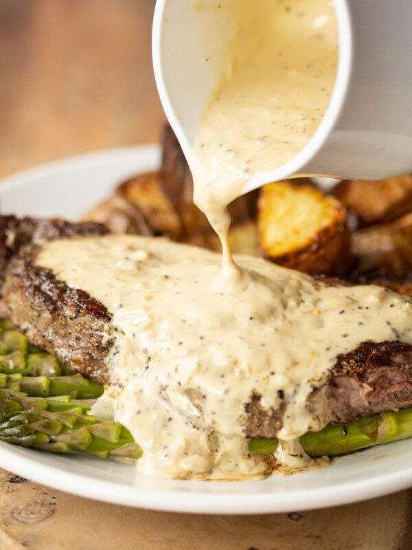 close up shot of small white pot pouring sauce over steak with potatoes and asparagus