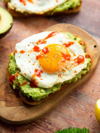 close up shot of avocado egg toast on small chopping board surrounded by garnish