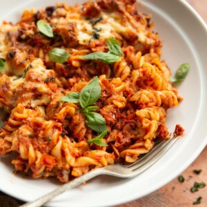 close up side shot of tomato tuna pasta bake in white bowl with silver fork garnished with basil