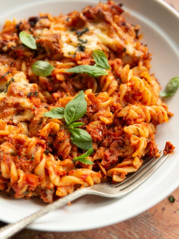 close up side shot of tomato tuna pasta bake in white bowl with silver fork garnished with basil