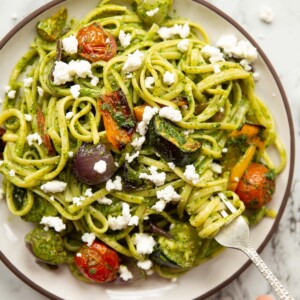 overhead shot of roasted vegetable pesto pasta on small white plate with hand twisting in silver fork