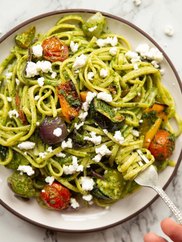 overhead shot of roasted vegetable pesto pasta on small white plate with hand twisting in silver fork