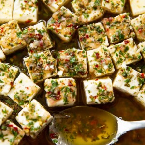 close up shot of marinated feta in glass dish with silver spoon