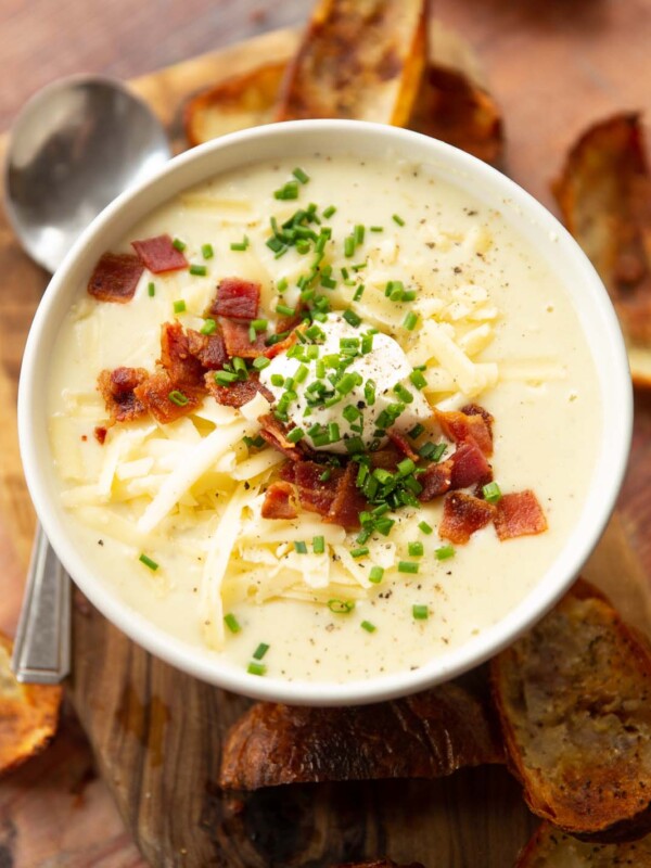 loaded baked potato soup in small white bowl on wooden chopping board surrounded by crispy potato skins