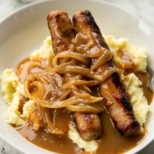 2 sausages on a bed of mashed potatoes with onion gravy in large white bowl