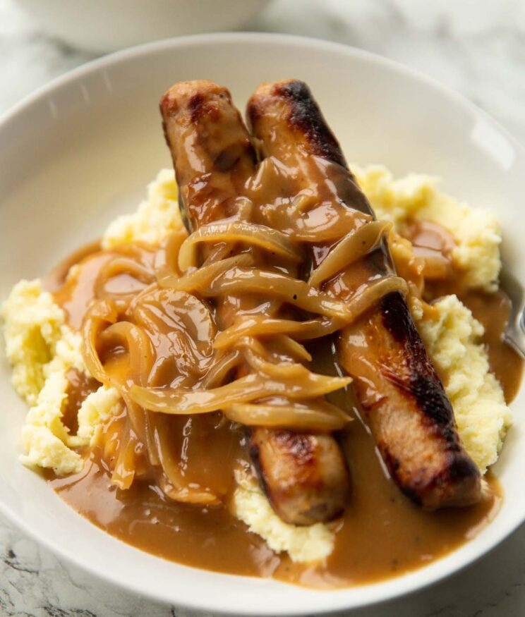 2 sausages on a bed of mashed potatoes with onion gravy in large white bowl
