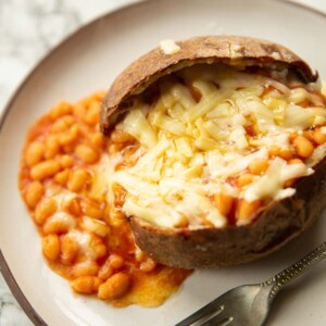 close up shot of jacket potato with beans and cheese on small white plate with silver fork to one side