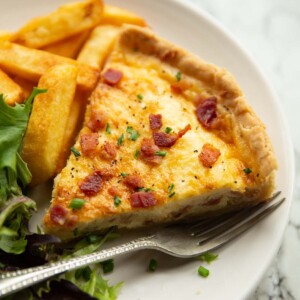 close up shot of slice of quiche lorraine on small white plate with chips and salad