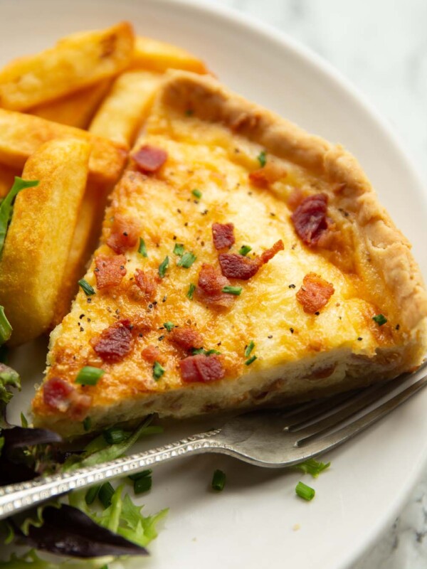 close up shot of slice of quiche lorraine on small white plate with chips and salad