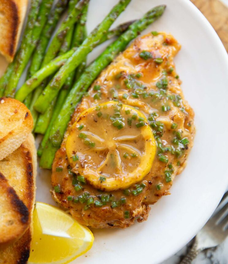 overhead shot of chicken francese on small white plate with bread and asparagus