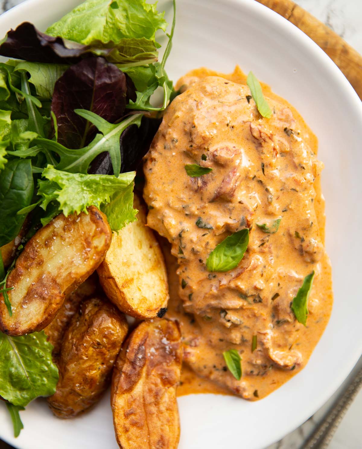 close up overhead shot of creamy sun dried tomato chicken breast on small white plate with potatoes and salad